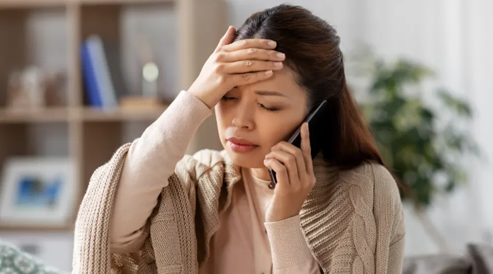 Concerned woman with a headache talking on the phone, possibly reporting mold-related health symptoms while sitting at home.