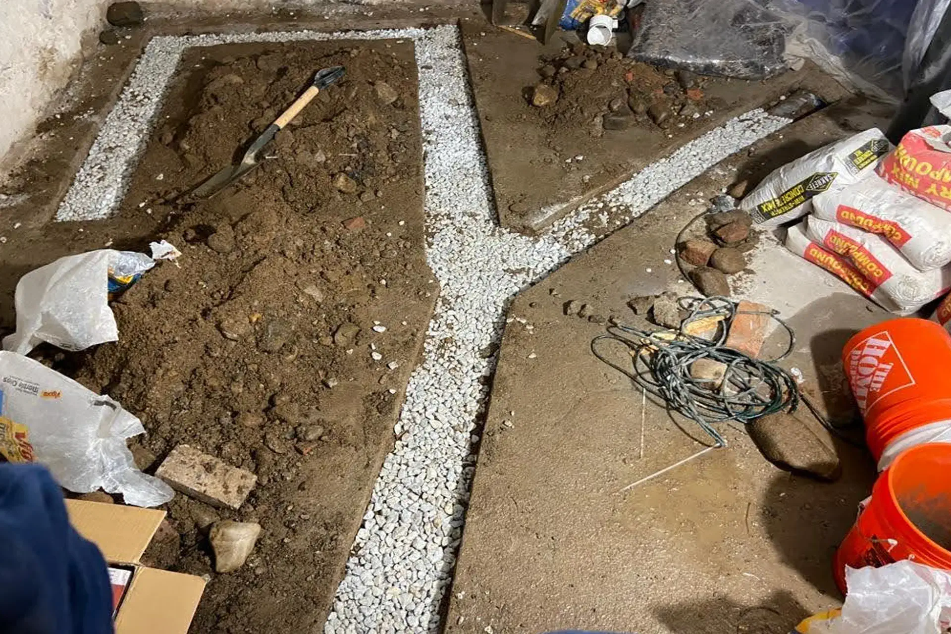Interior basement trenching showing freshly excavated channels in a Y-shaped pattern, with gravel placed for a new French drain system, surrounded by soil, tools, and construction materials during a drainage installation project.
